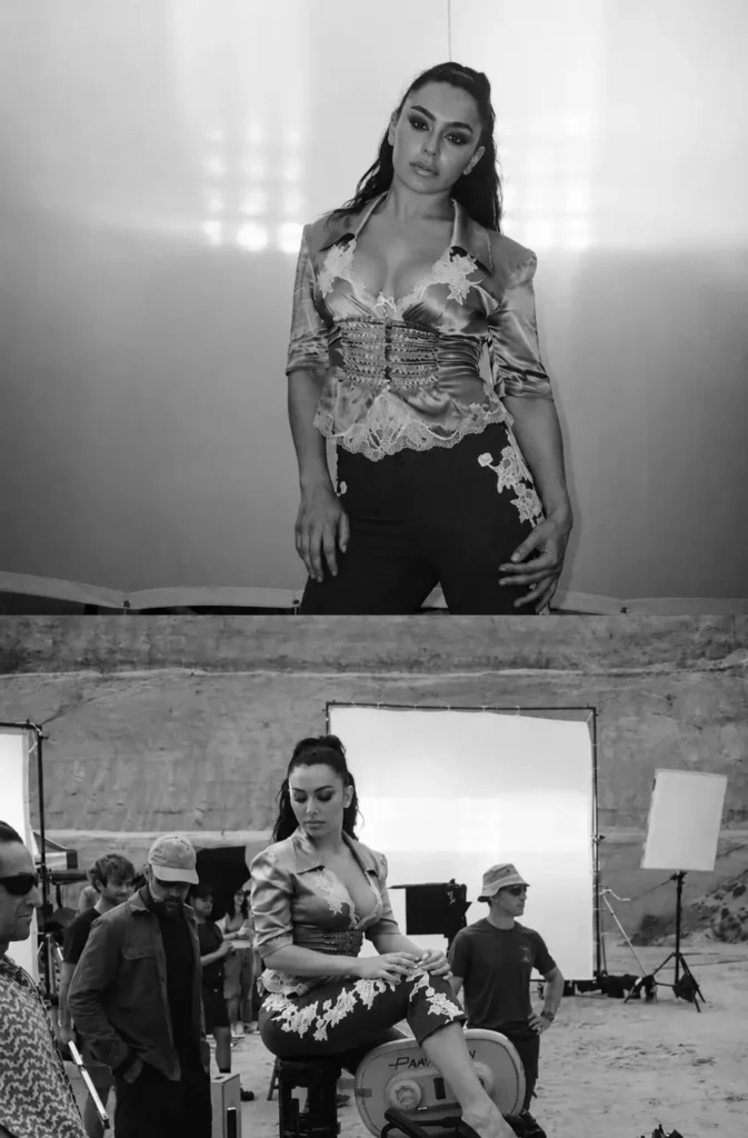 Black-and-white behind-the-scenes photo of Charli xcx seated on film equipment at an outdoor production set, surrounded by crew members, lighting rigs, and camera gear