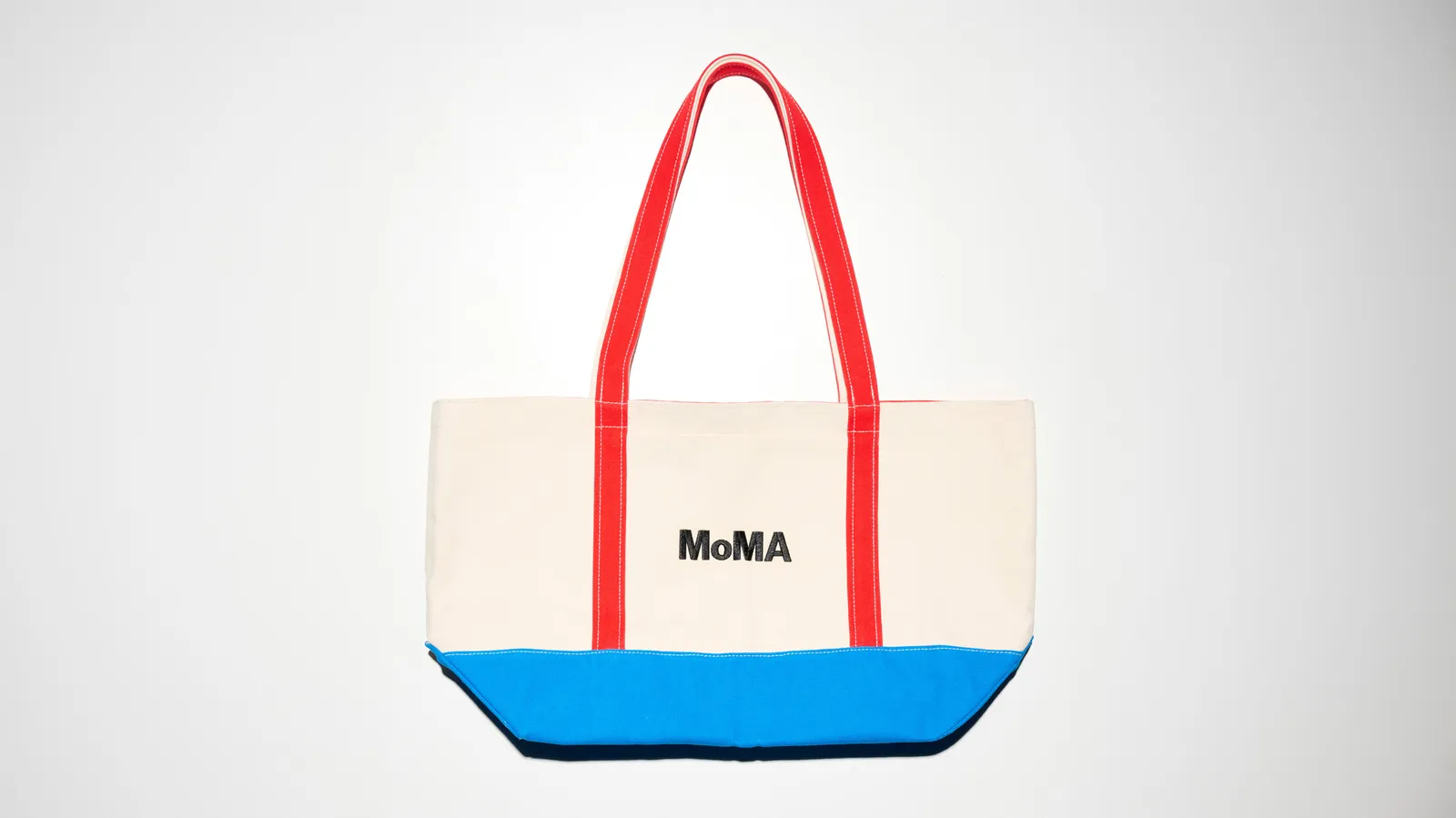 Cream canvas MoMA tote bag featuring bright red shoulder straps, a vivid blue reinforced base, and a centered black MoMA logo, photographed against a clean white studio background.