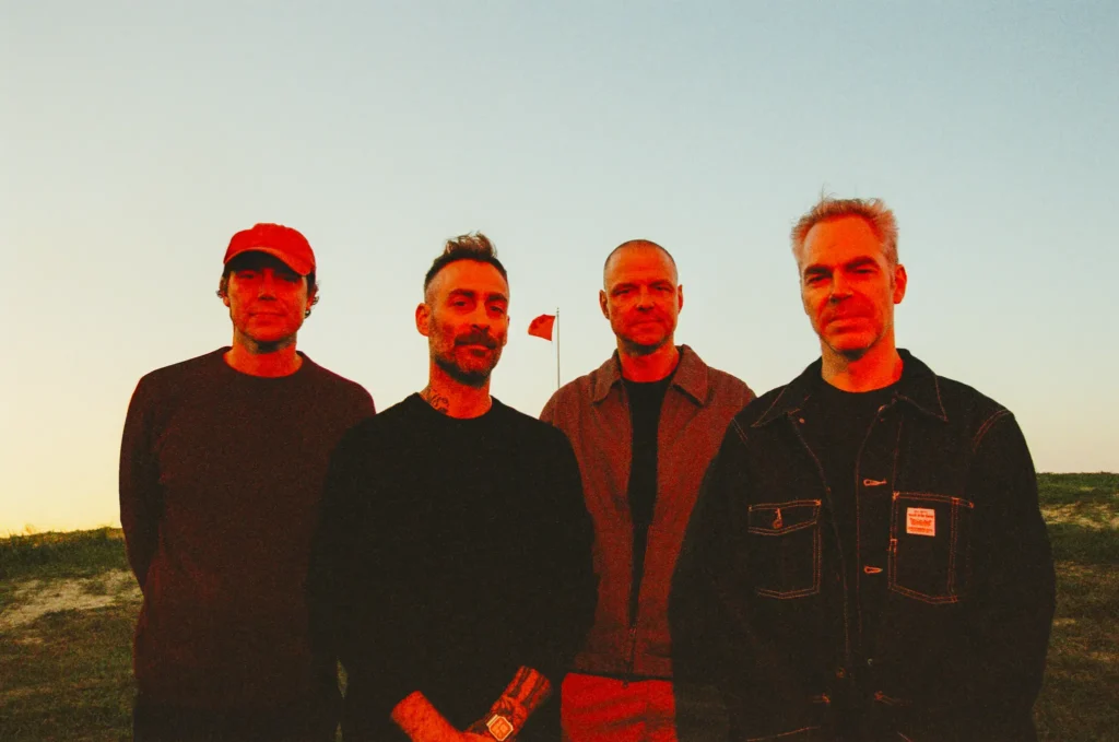 Four band members stand outdoors at dusk beneath a pale evening sky, facing the camera with a red flag view in the background
