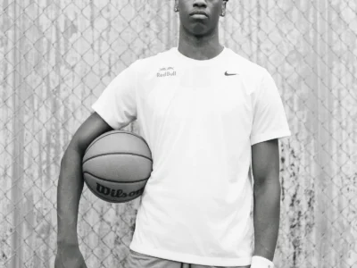 Black-and-white portrait of a basketball player AJ Dybantsa holding a Wilson basketball while standing in front of a chain-link fence and corrugated metal wall, wearing a white Nike T-shirt and gray shorts