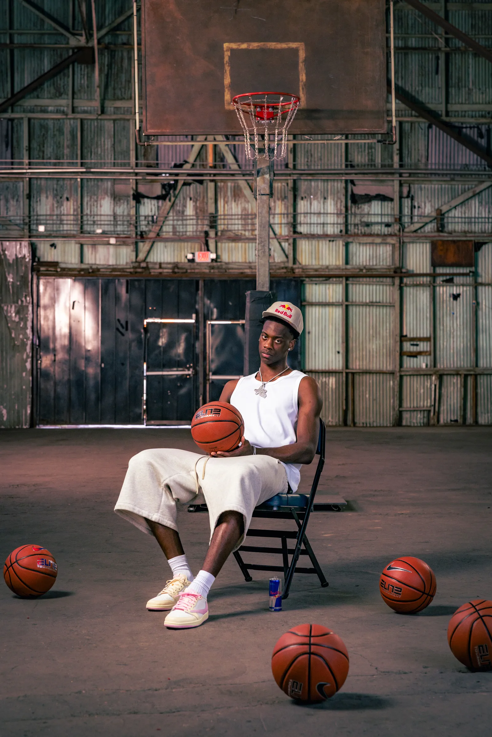Basketball player seated beneath a hoop in an industrial gym, holding a basketball with several balls scattered across the floor.