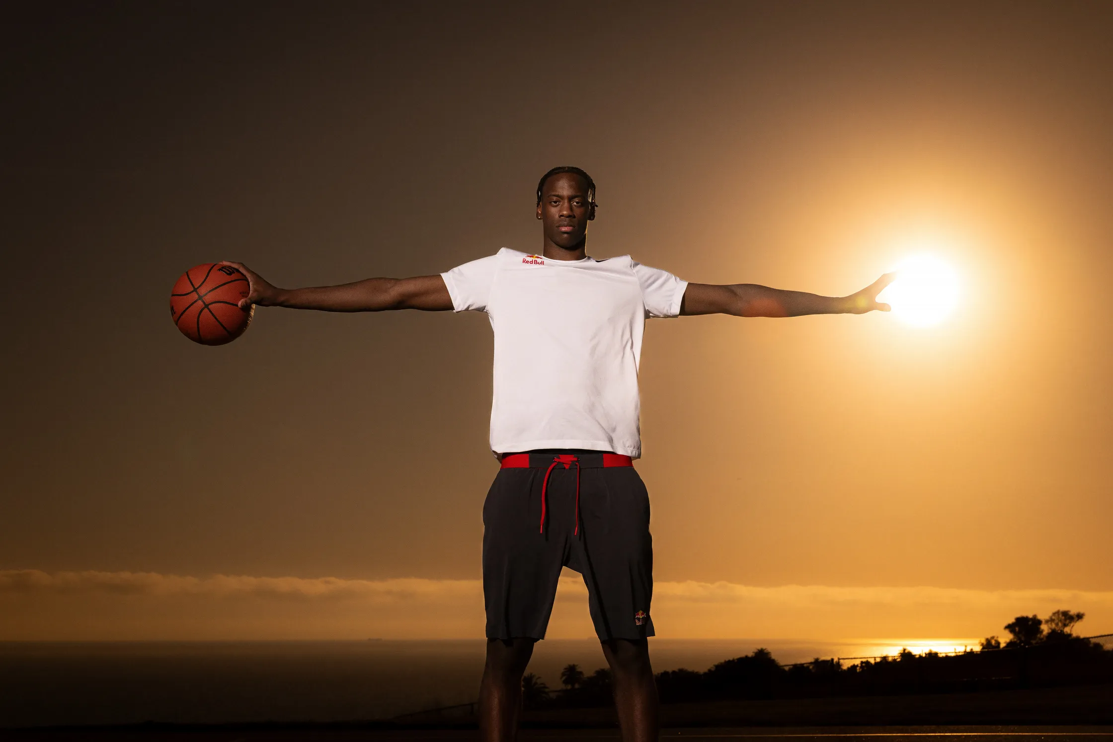Basketball player balancing a basketball with outstretched arms against a dramatic sunset over the ocean.