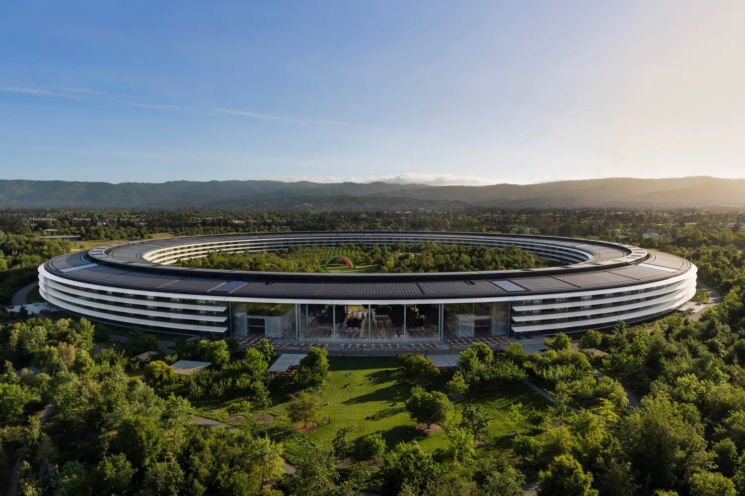 Aerial view of Apple Park in Cupertino, California, showing the company's iconic circular headquarters surrounded by landscaped gardens, with the central courtyard and distant hills under a clear blue sky.