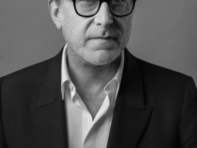 A black-and-white studio portrait of Romain Spitzer wearing black rectangular eyeglasses, a dark tailored blazer, and an open-collar white dress shirt. The subject faces the camera with a calm, serious expression against a plain gray background, creating a clean, professional editorial look