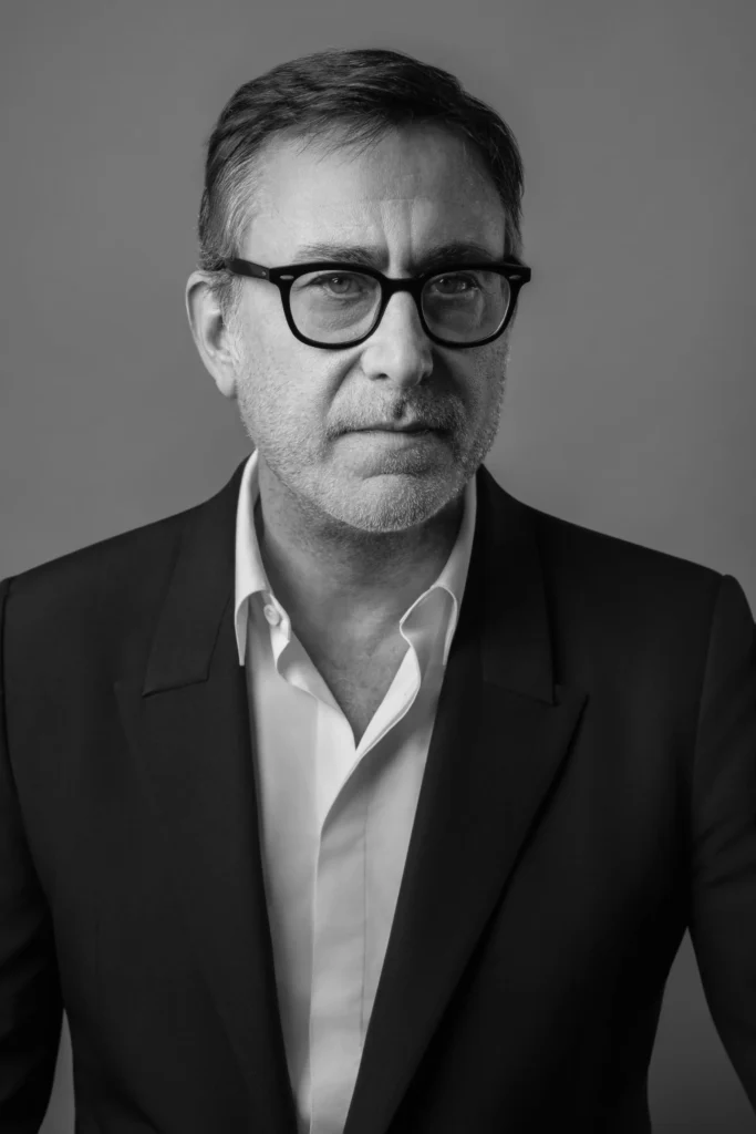 A black-and-white studio portrait of Romain Spitzer wearing black rectangular eyeglasses, a dark tailored blazer, and an open-collar white dress shirt. The subject faces the camera with a calm, serious expression against a plain gray background, creating a clean, professional editorial look