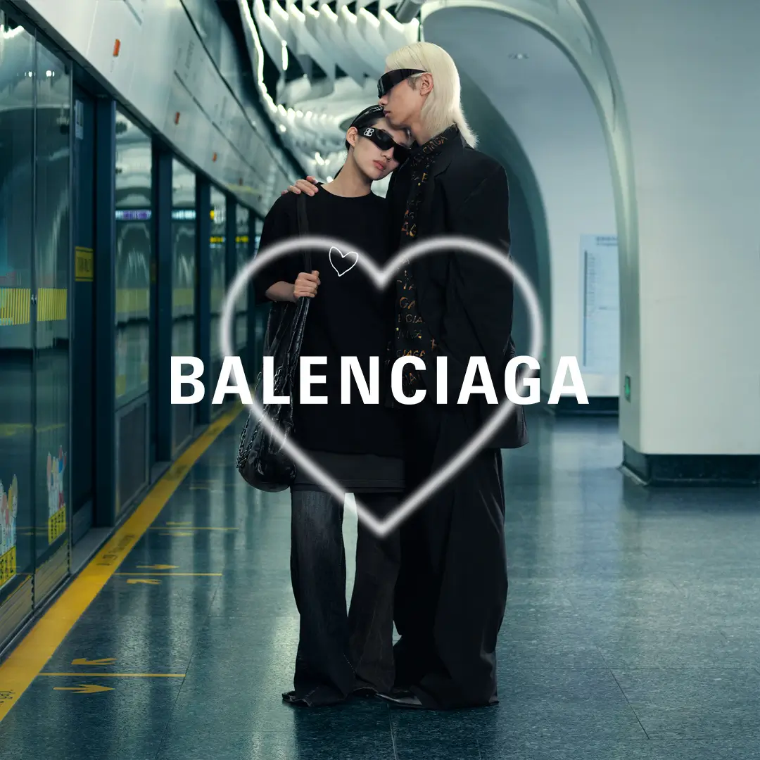 Two models dressed in oversized black Balenciaga looks embrace on a modern subway platform while wearing futuristic wraparound sunglasses. A glowing hand-drawn heart graphic frames the pair with the Balenciaga logo centered across the image, reinforcing the romantic theme of the QIXI 26 campaign.