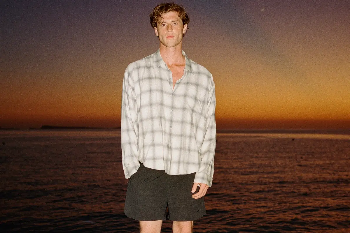Male model wearing a light gray plaid button-up shirt, black shorts, and brown suede loafers while standing on a rocky shoreline at sunset with calm ocean waters and a glowing orange sky.