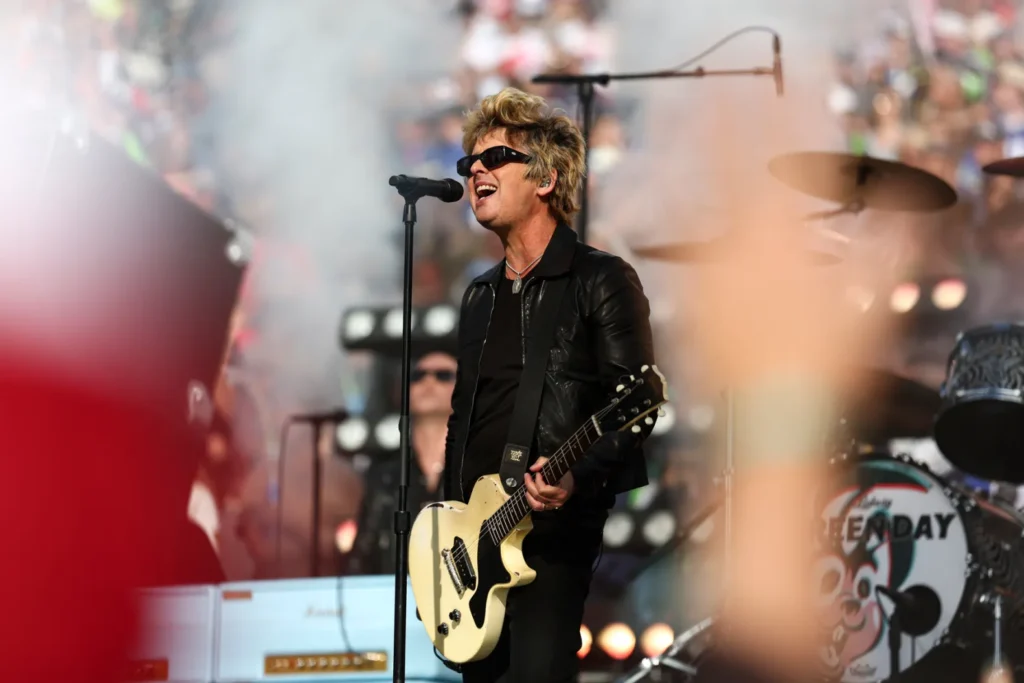 Green Day frontman Billie Joe Armstrong performs on an outdoor stage, singing into a microphone while playing a cream-colored electric guitar. Dressed in a black leather jacket and sunglasses, he stands amid drifting stage smoke with the band's drum kit behind him and a packed festival crowd filling the background. Blurred audience members in the foreground frame the energetic live show