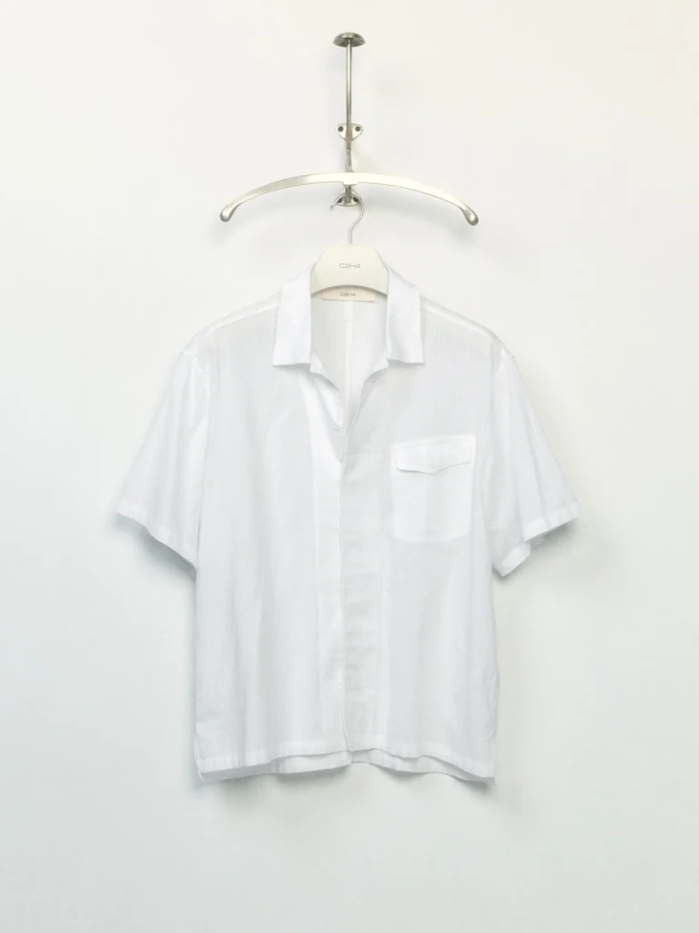 White short-sleeve camp-collar shirt with a button front, single flap chest pocket, and lightweight fabric hanging on a rack