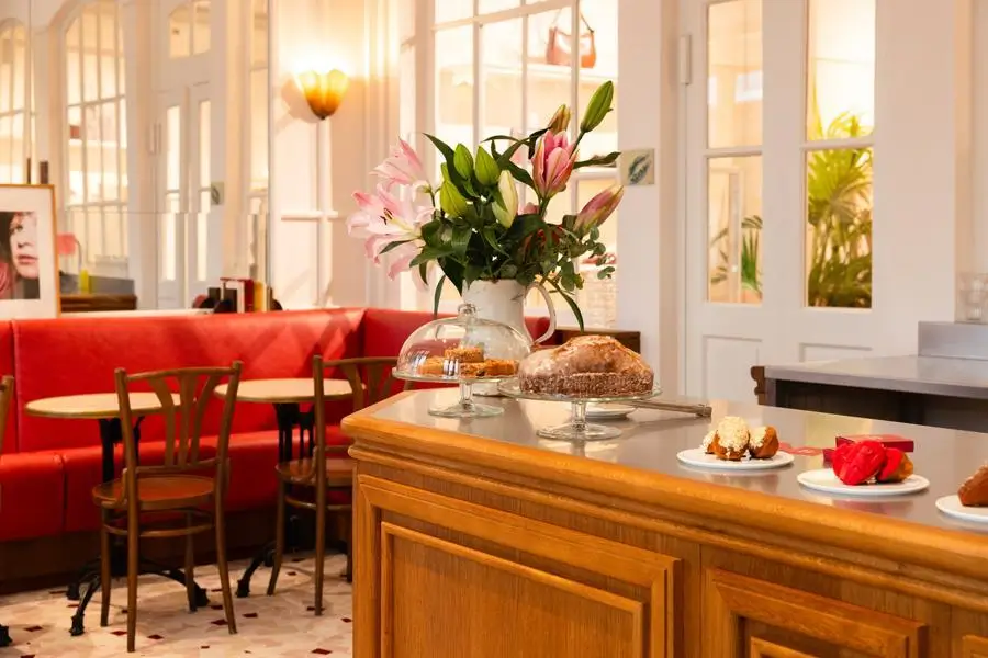 A warm café interior with red banquette seating, wooden tables and chairs, a marble counter displaying fresh pastries and flowers, and soft natural light streaming through tall windows.