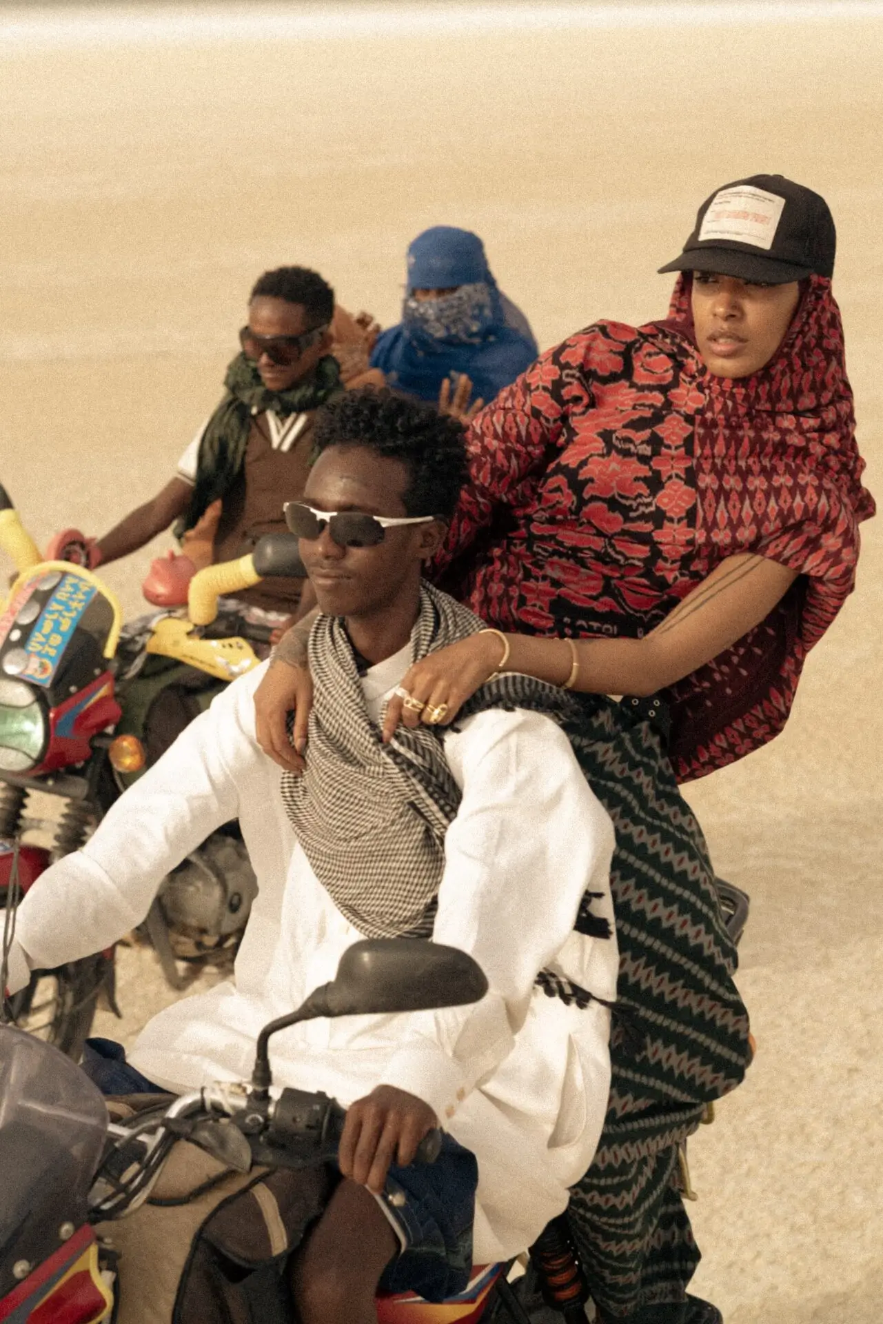 Fashion editorial featuring two people riding a motorcycle across a sunlit salt flat, wearing patterned scarves, layered garments, sunglasses, and caps, with additional riders following in the background.