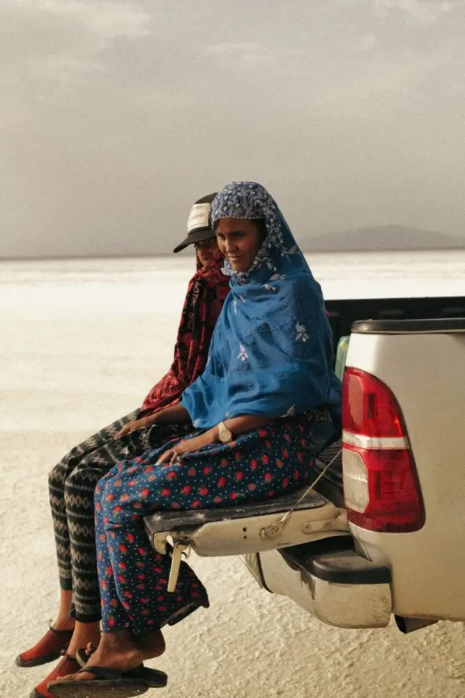 Two people sit on the open tailgate of a pickup truck overlooking a bright salt flat, dressed in colorful patterned wraps and traditional-inspired clothing under warm afternoon light.