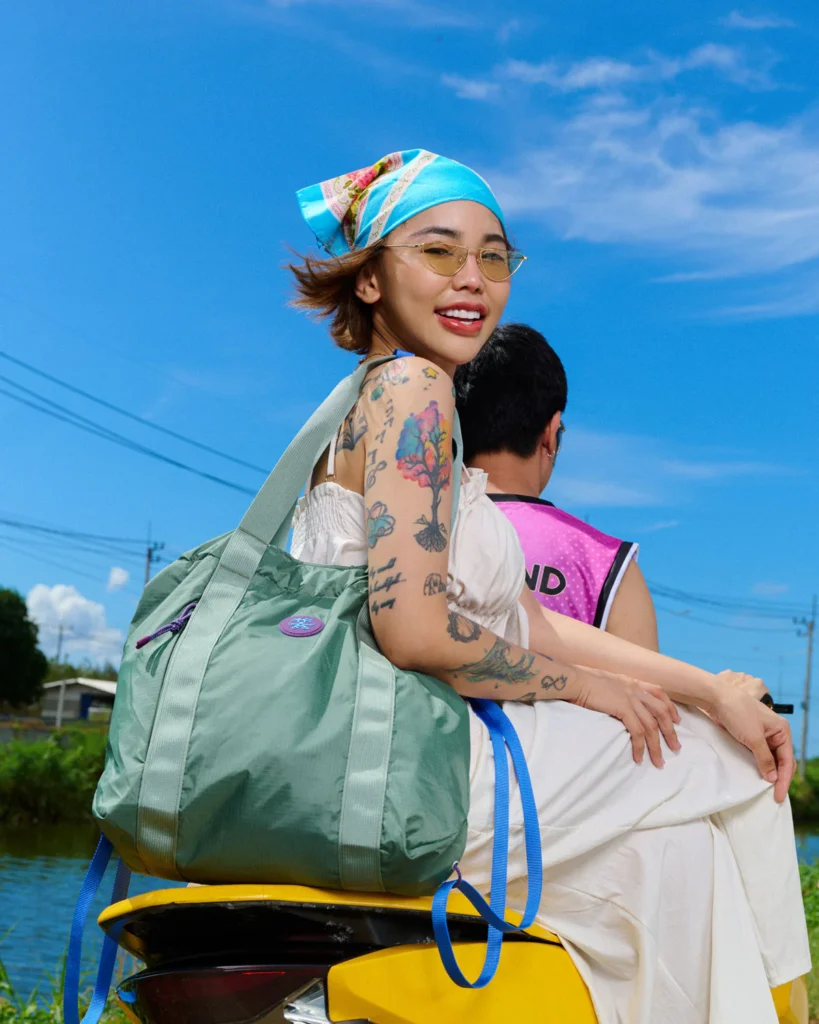 Person wearing a large sage green Crumpler shoulder tote while seated outdoors under a bright blue sky, showcasing the spacious everyday carry bag in a lifestyle set