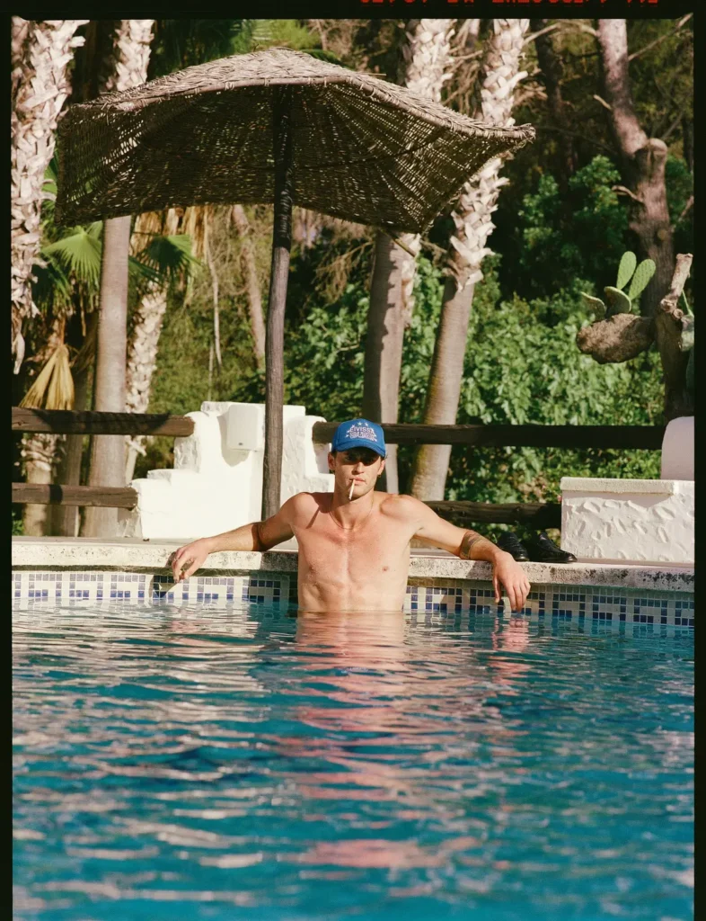 Male model wearing a blue trucker cap lounges shirtless in a resort swimming pool beneath a woven parasol, surrounded by palm trees and Mediterranean-inspired architecture