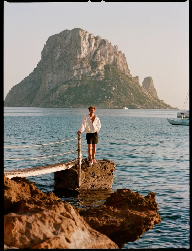 Male model wearing a white zip hoodie, black shorts, and white sneakers with pink laces stands on a small stone pier overlooking turquoise water and a dramatic rocky island