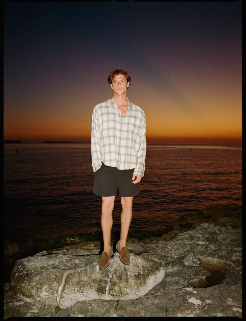 Wide view of male model standing on a stone pier in a white hoodie, black shorts, and white sneakers with pink laces against a calm Mediterranean bay and towering island backdrop