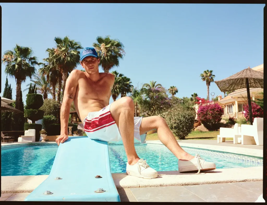Shirtless male model wearing white athletic shorts, white platform sneakers, and a blue trucker cap sits beside a resort swimming pool lined with palm trees and flowering garden