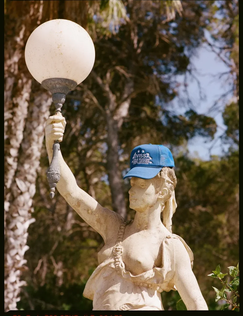 Stone garden statue holding a vintage lamp wears a blue mesh trucker cap embroidered with white lettering in a play outdoor fashion install