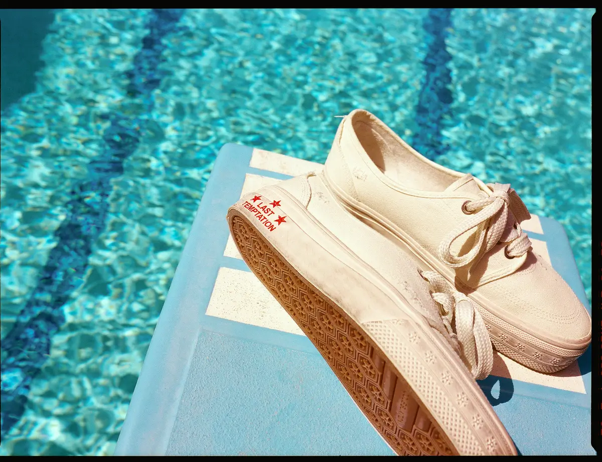 Cream canvas sneakers with textured rubber soles resting on a blue pool slide beside a sparkling turquoise swimming pool, highlighting the casual summer footwear in bright natural sunlight.