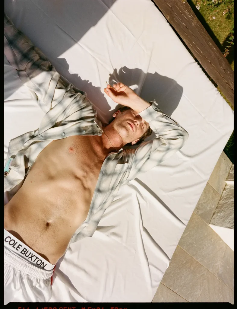 Male model reclines on a white outdoor daybed wearing an open plaid shirt and white athletic shorts, relaxing in bright Mediterranean sunshine