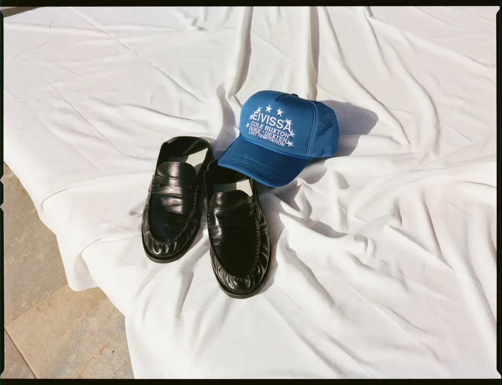 Black leather penny loafers and a blue embroidered trucker cap arranged on a white outdoor cushion in natural sunlight