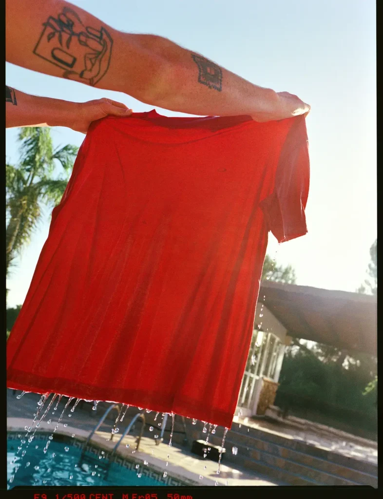 Hands hold up a soaked bright red athletic Tee dripping water above a swimming pool with sunlight shining through the lightweight fabric