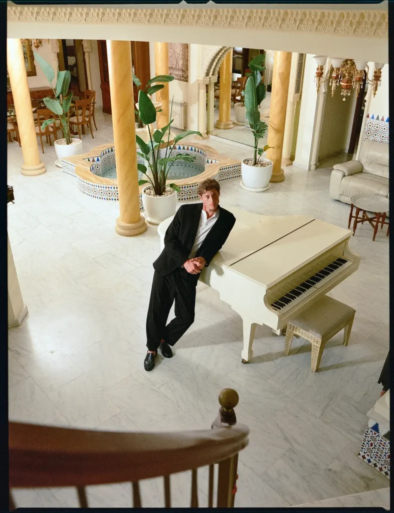 Male model dressed in a black suit and white shirt leans against a white grand piano inside an elegant atrium with marble floors and decorative column