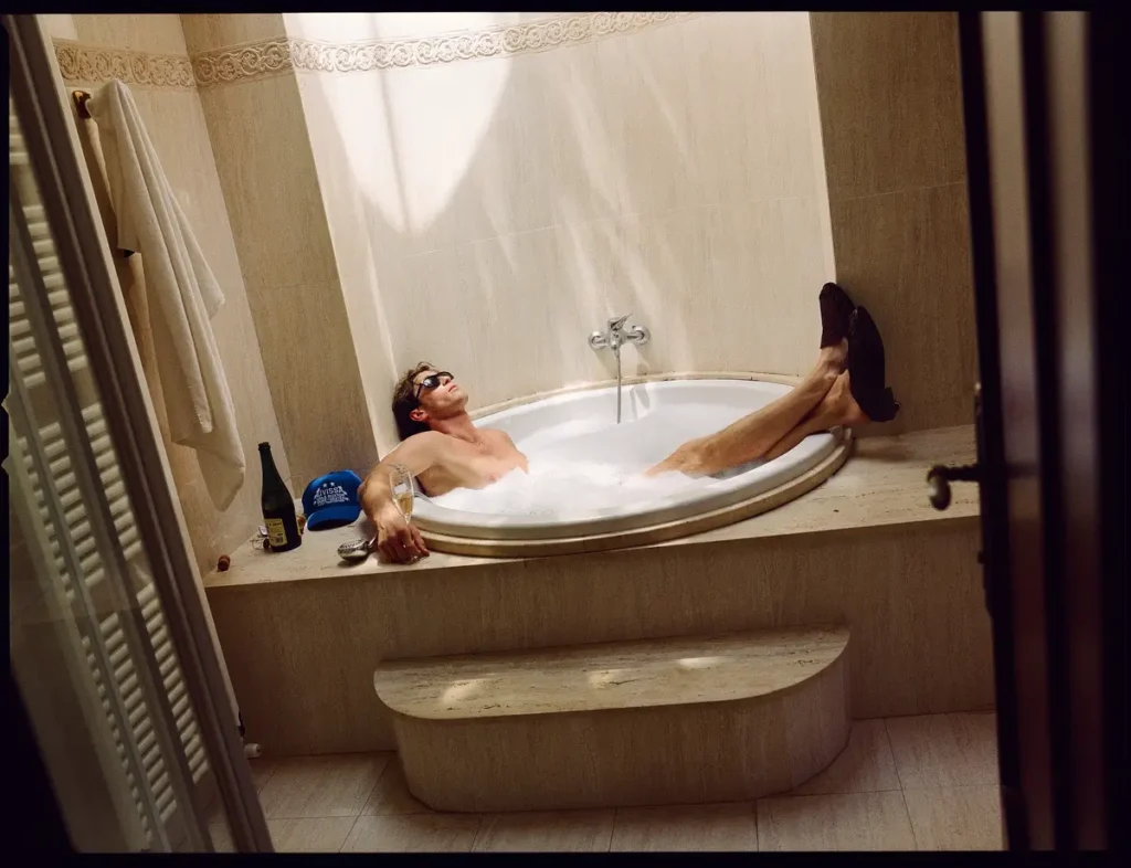Male model relaxes in a bubble bath wearing sunglasses and brown suede loafers while holding a champagne flute beside a bottle and blue trucker cap