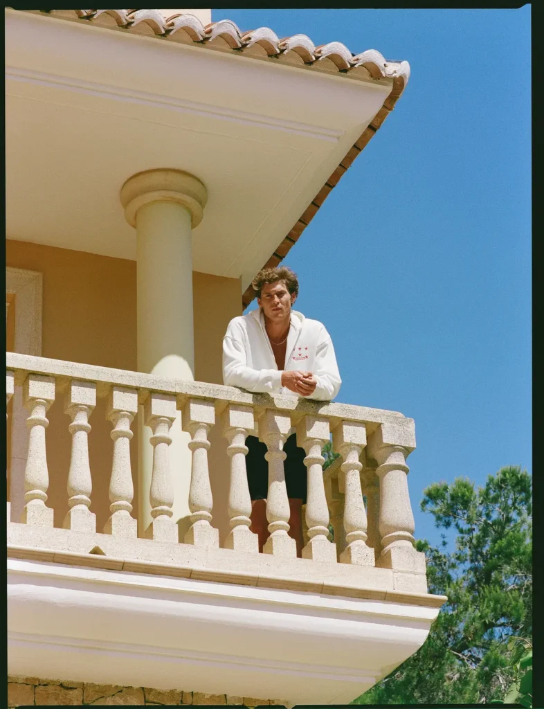 Male model wearing a white zip hoodie and black shorts leans on a stone balcony beneath a clear blue sky at a Mediterranean-style villa