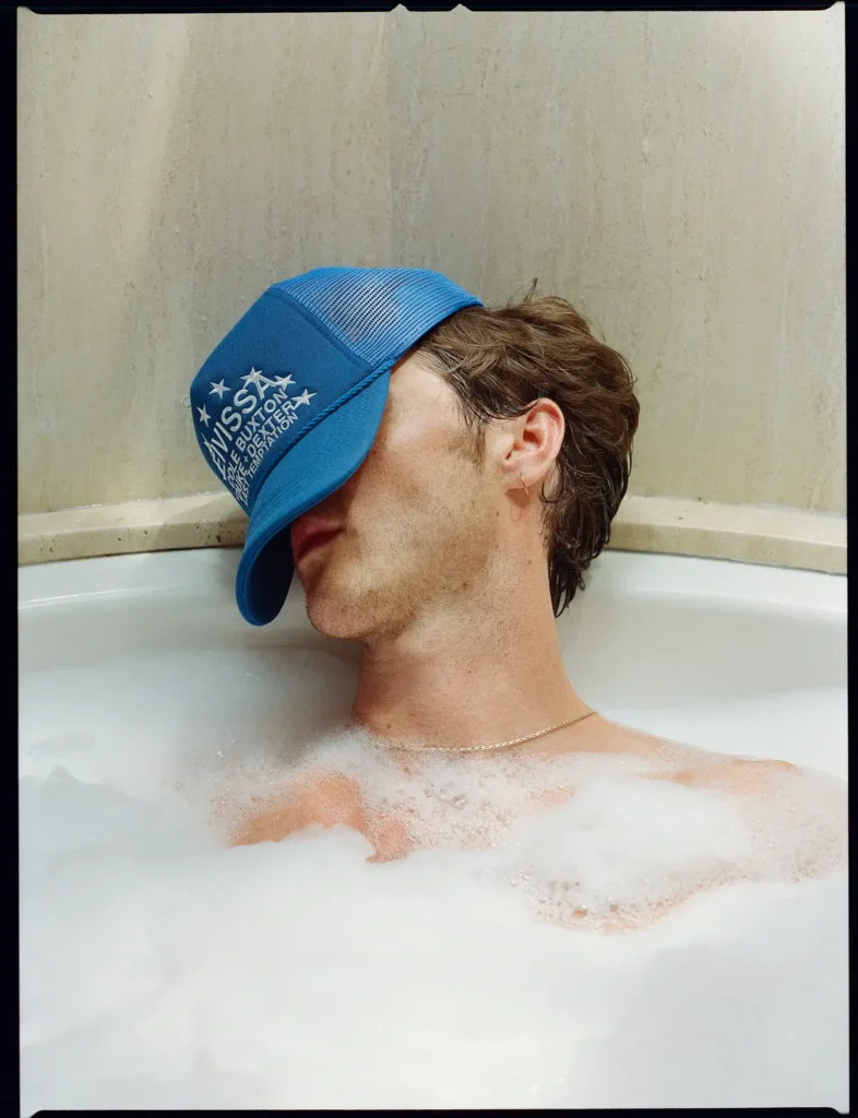 Close-up portrait of a male model relaxing in a bubble bath wearing a blue embroidered trucker cap pulled low over his face and a thin gold necklace