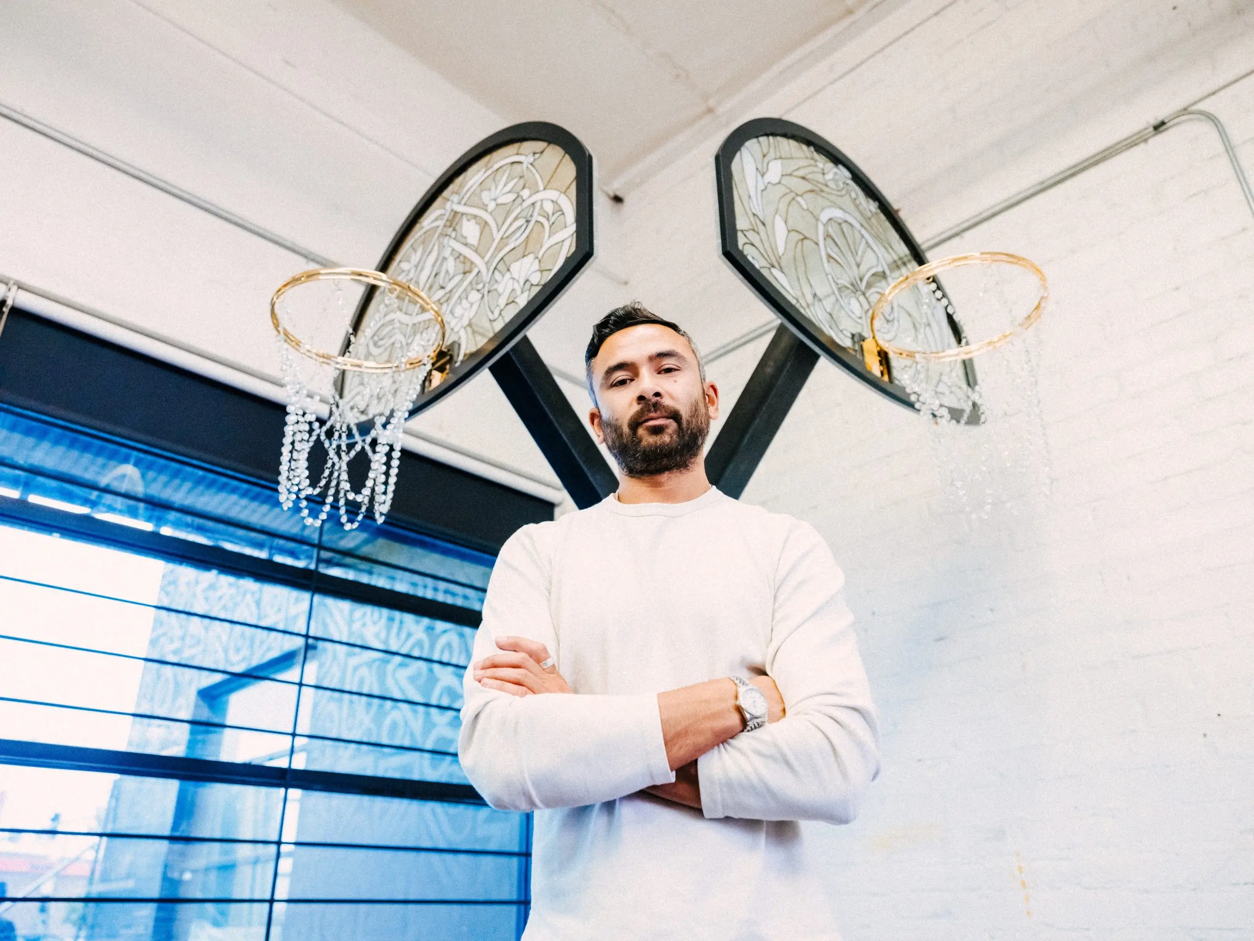 Portrait of artist Victor Solomon standing with folded arms beneath a pair of his signature sculptural basketball hoops, featuring ornate backboards and chain nets inside a bright industrial studio.