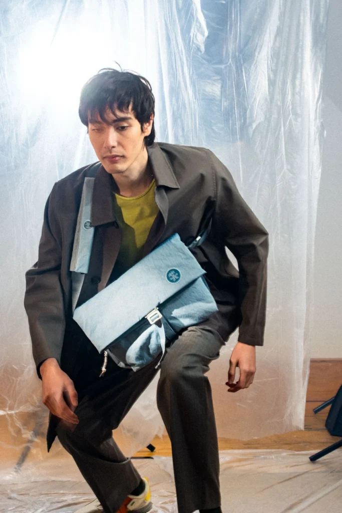 Model wearing a light blue Crumpler messenger bag across the body inside a studio with translucent plastic backdrops, emphasizing the oversized everyday carry sil