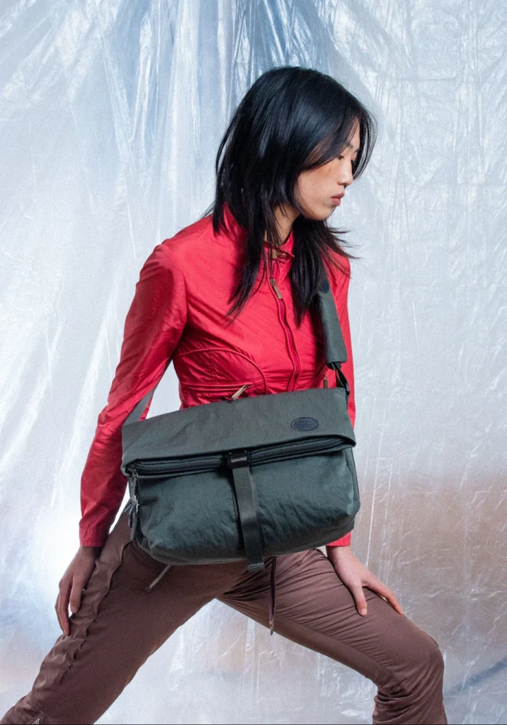 Model wearing a dark green Crumpler shoulder bag styled with a bright red jacket in a studio setting, highlighting the bag's folded top design and oversized capacity