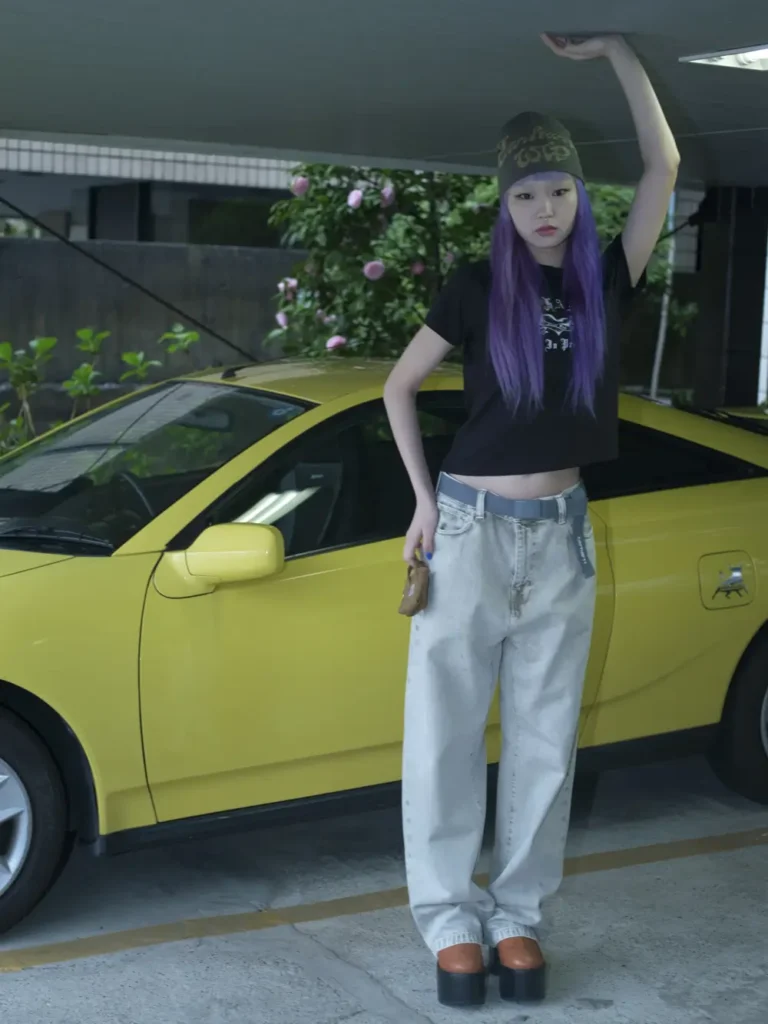 Model with long purple hair stands beside a yellow sports car wearing a graphic beanie, black cropped T-shirt, faded baggy jeans, platform shoes, and a gray belt in an urban parking garage