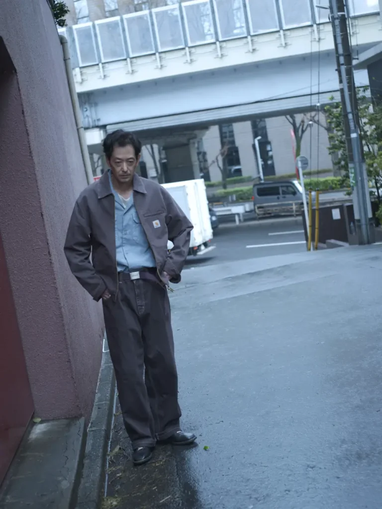 Person wearing a gray work jacket over a light blue button-up shirt and matching work pants stands on a wet city sidewalk beside a building, creating a rugged utility-inspired look