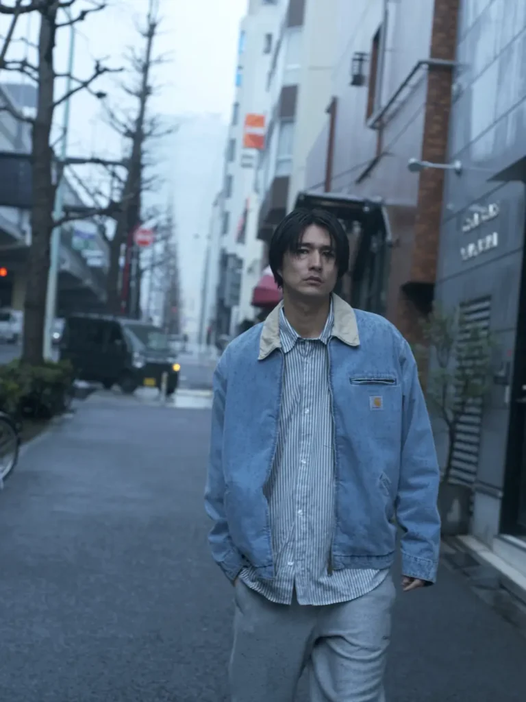 Model walks through a quiet urban street wearing a light blue washed work jacket layered over a striped button-up shirt with gray sweatpants, showcasing relaxed day workwear style