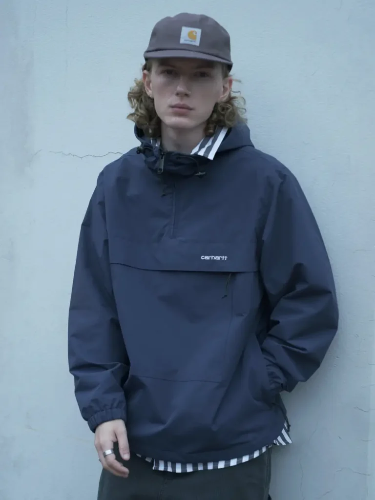 Portrait of a model wearing a navy Carhartt WIP hooded anorak layered over a striped shirt and paired with a brown Carhartt cap against a minimalist blue wall