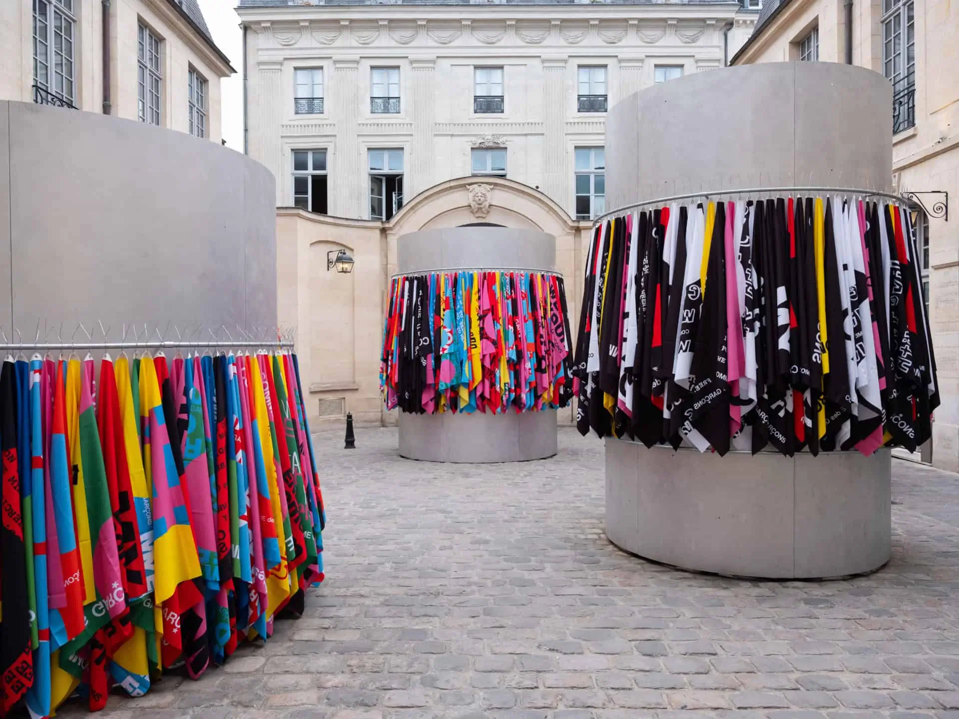Colorful flag installation transforms a historic Paris courtyard with cylindrical displays wrapped in vibrant graphic textiles against elegant stone architecture.