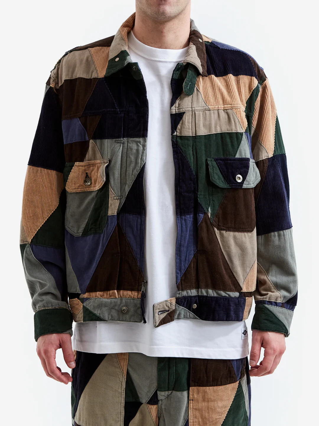 Model wearing a multicolor patchwork trucker jacket crafted from mixed corduroy and cotton panels in olive, navy, brown, tan, and gray, featuring geometric triangular panels, flap chest pockets, and a relaxed workwear-inspired fit.