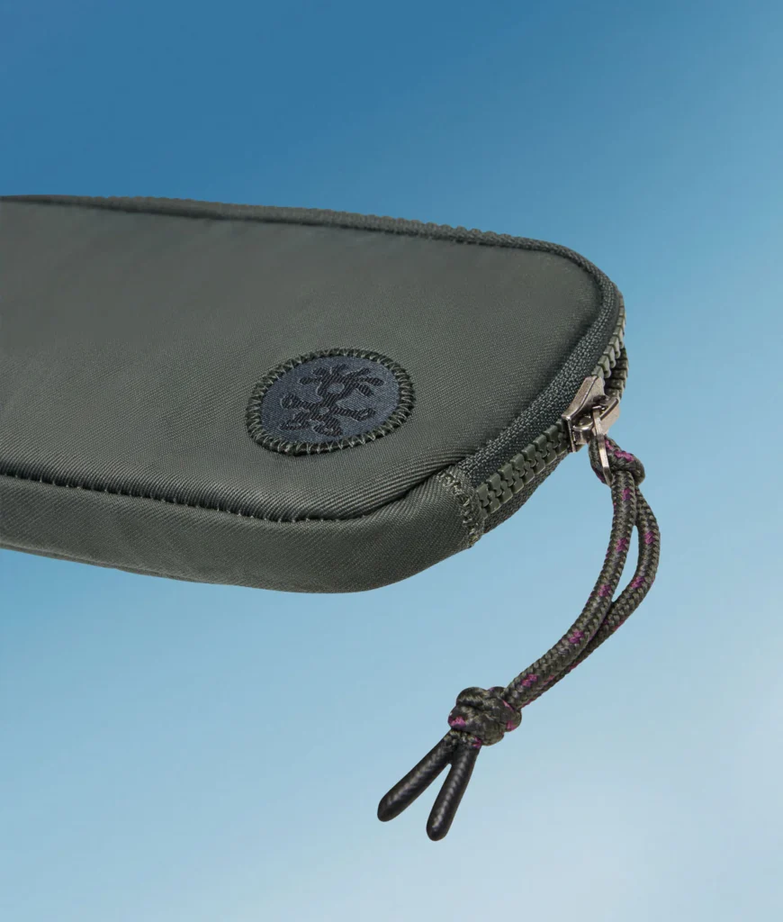 Close-up of a dark green Crumpler zip pouch highlighting the circular stitched logo, heavy-duty zipper, and knotted cord zipper pull against a blue background