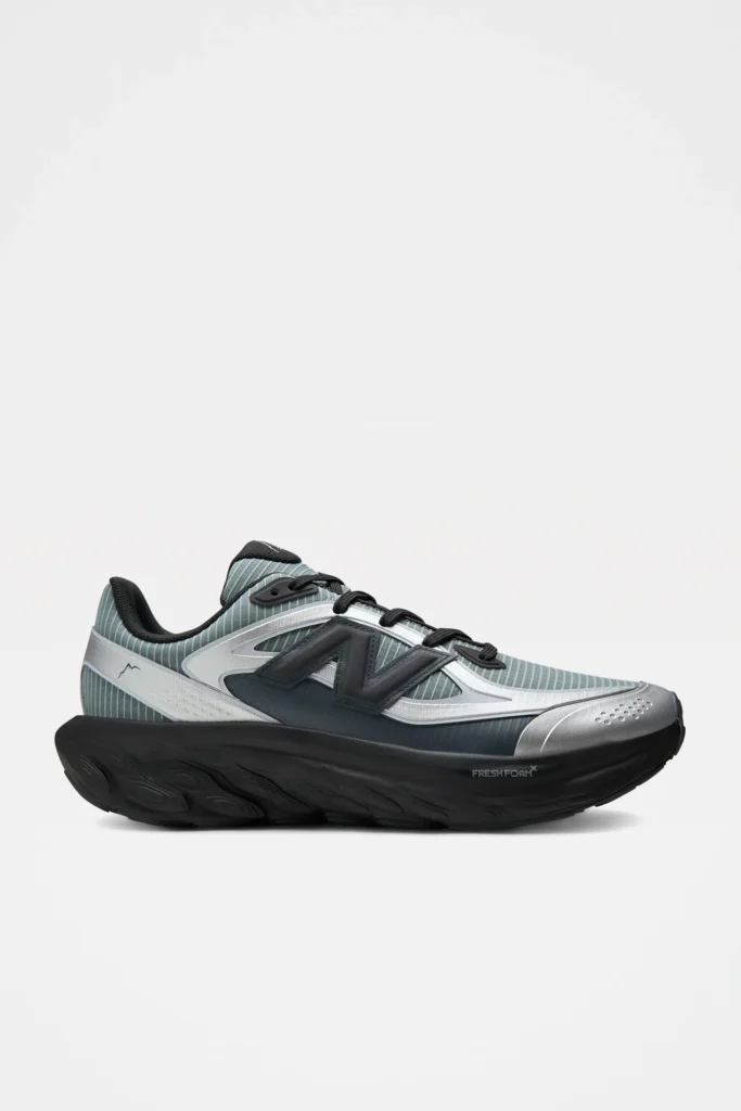 Side profile of a New Balance running shoe featuring a muted sage-gray striped mesh upper, metallic silver overlays, oversized black "N" logo, black Fresh Foam X midsole, black laces, and a clean white studio background