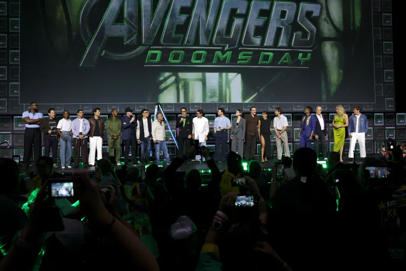 The cast of Marvel Studios' Avengers: Doomsday stands together onstage during the Hall H presentation at San Diego Comic-Con, gathered beneath a massive Avengers: Doomsday logo as fans photograph the surprise appearance. The wide stage reveal marked one of Marvel's biggest announcements for the upcoming crossover film.