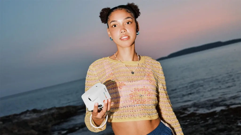 Person standing along the shoreline at dusk holding the white Polaroid Flip instant camera, with calm ocean waters and a colorful sunset sky creating a relaxed coastal backdrop