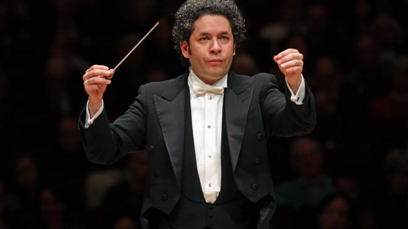 Orchestra conductor in a black tailcoat and white bow tie leading a performance with a raised baton before a blurred concert hall audience.