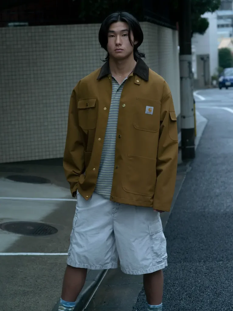 Model wearing a brown Carhartt WIP chore jacket over a striped knit polo with oversized white cargo shorts, socks, and sneakers stands on a rain-soaked residential street