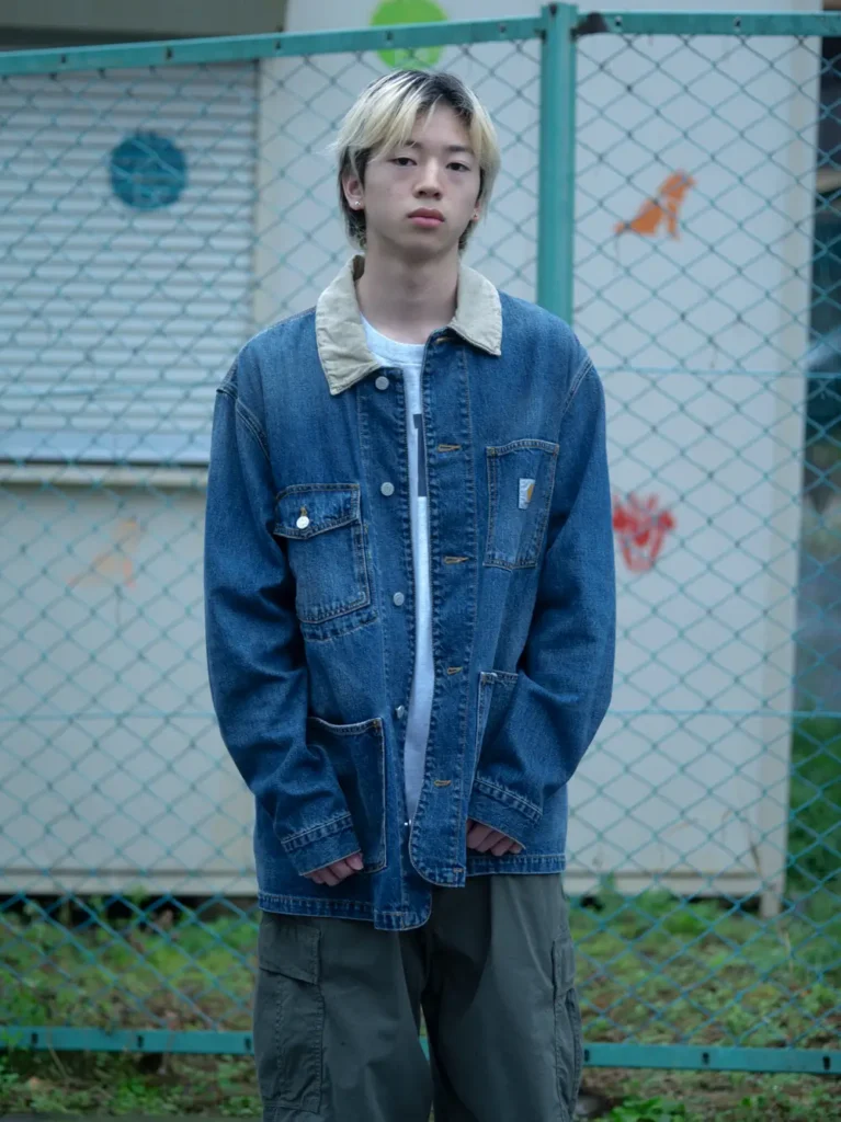 Model in an oversized blue denim chore jacket with a corduroy collar, white T-shirt, and olive cargo pants poses in front of a chain-link fence for a casual streetwear look