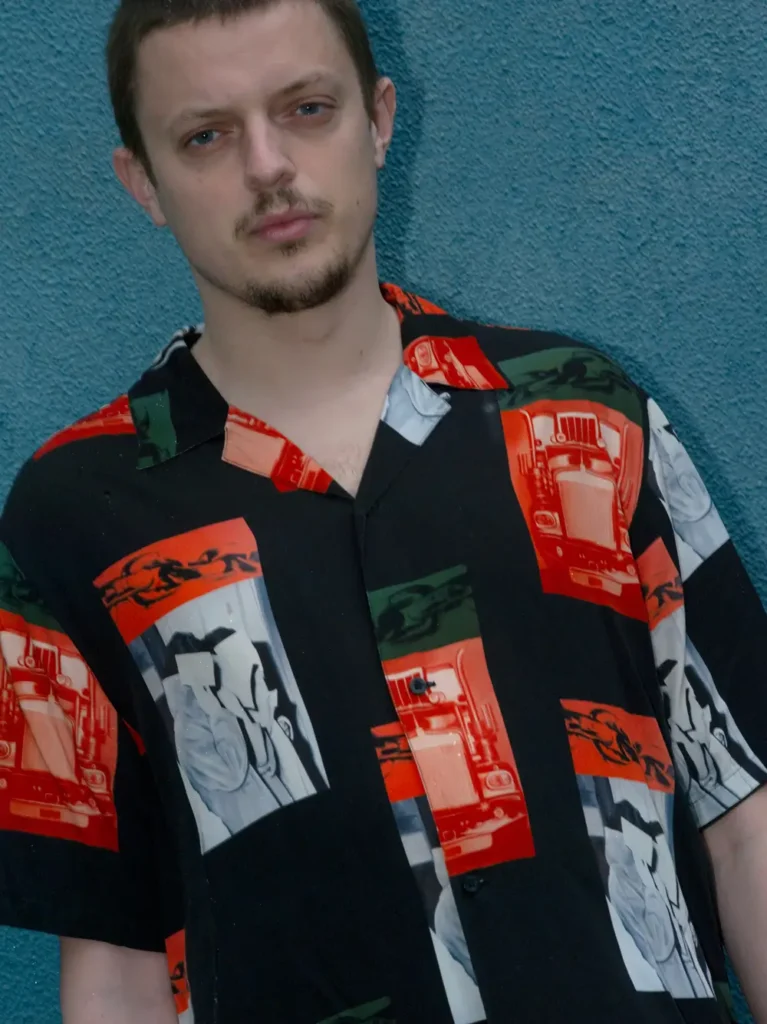 Portrait of a model wearing a black short-sleeve button-up shirt featuring bold red, white, and green graphic artwork against a textured blue wall
