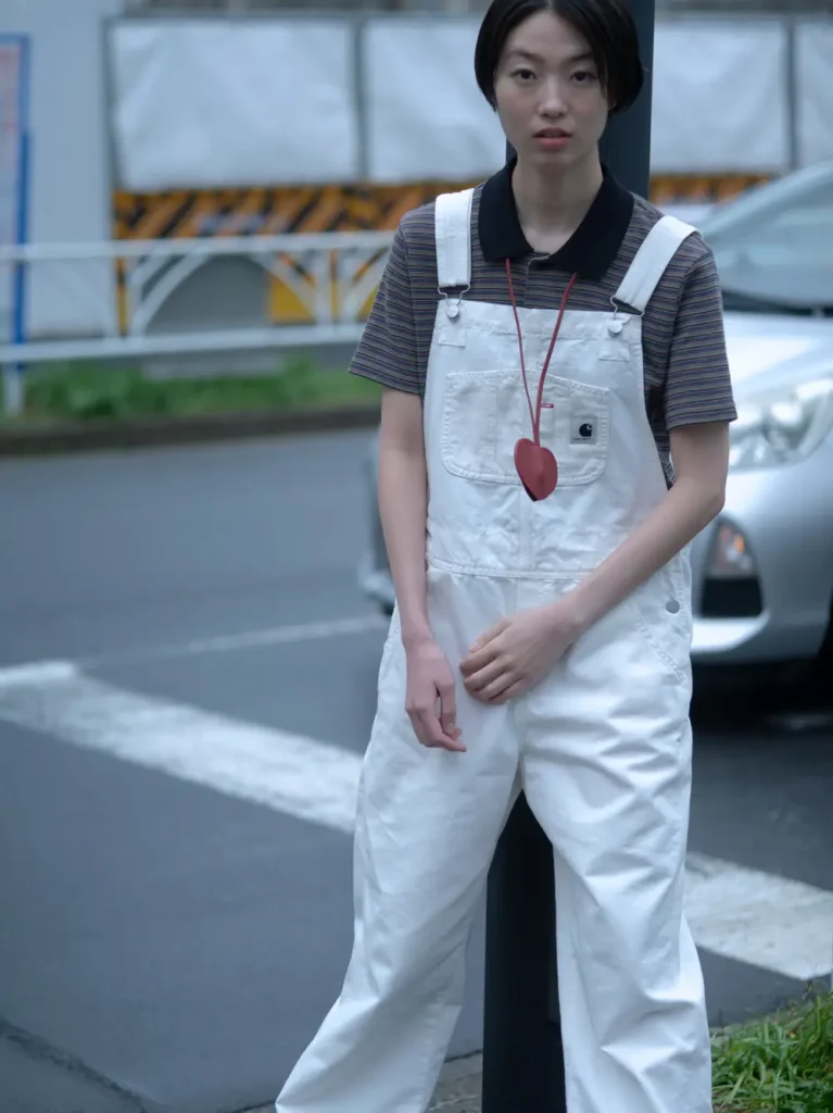 Model wears white Carhartt WIP overalls layered over a striped polo shirt with a large red heart pendant while standing beside a city street and traffic lane