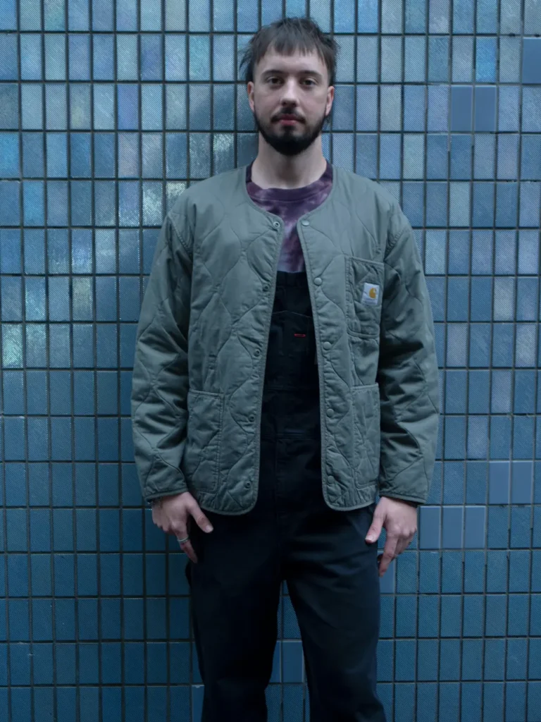 Portrait of a model wearing an olive quilted liner jacket over a tie-dye T-shirt and black overalls, standing against a tiled wall with a clean workwear-inspired show