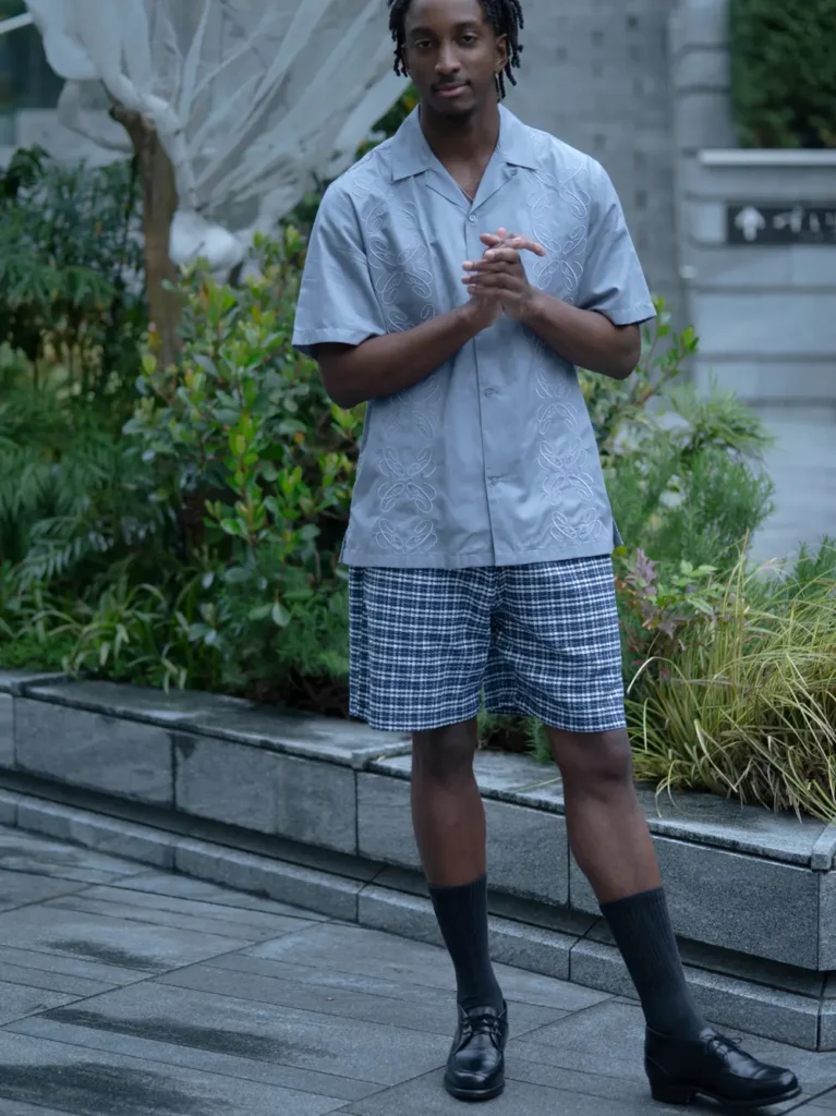 Model poses in a light gray embroidered camp-collar shirt, blue checkered shorts, black crew socks, and leather loafers in a landscaped urban set